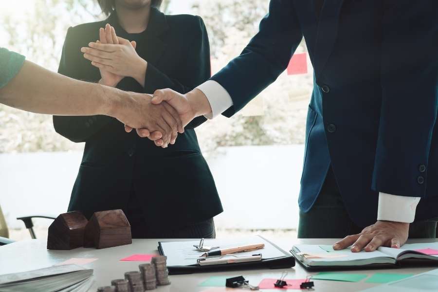 Une poignée de main professionnelle au-dessus d'une table de bureau avec des modèles de maison et de l'argent, symbolisant une transaction immobilière réussie dans un quartier où investir.