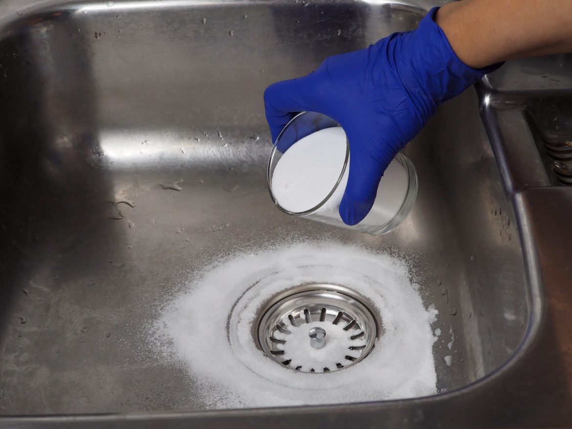 Une main avec un gant bleu verse du bicarbonate de soude autour de la bonde d'un évier en inox pour entretenir sa plomberie naturellement