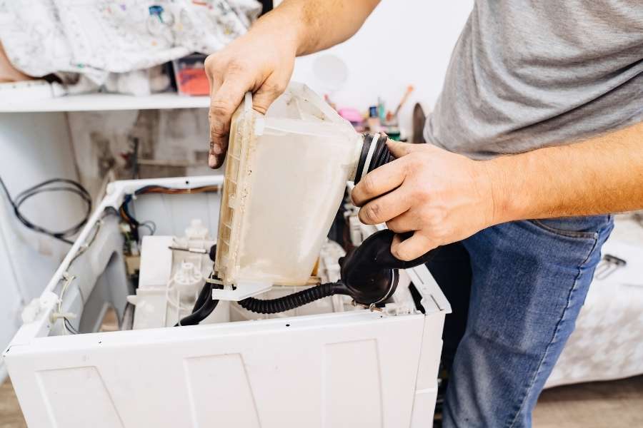 Un technicien manipule le bac à produits et les pièces détachées à l'intérieur d'un lave-linge ouvert.